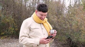 young man wearing yellow scarf enjoying autumn days breathing fresh air, enjoying walking in the park, drinking from thermos and using smartphone video