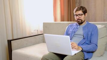 Relaxed serious millennial guy student freelancer using laptop device leaning on sofa at home office, focused mixed race entrepreneur working distantly typing on notebook in apartment video