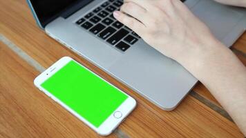 A woman is typing on a laptop with a green screen, with a mobile phone beside her, ideal for app design, digital content creation, and product mockup presentations. video