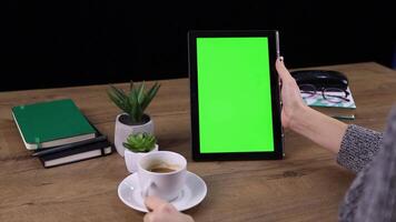 A woman holding a tablet with a green screen while enjoying a cup of coffee, sitting in a cozy environment. video