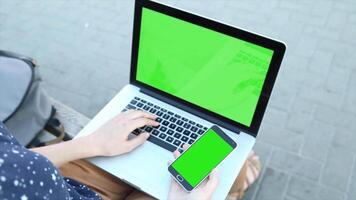 A person sitting on a bench using a laptop and mobile phone with green screens, perfect for app design, digital content, and product mockup presentations. video