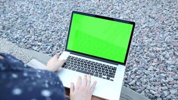 A person sitting on a bench outdoors using a laptop computer with a green screen, ideal for digital content creation, app design, and mockup presentations. video