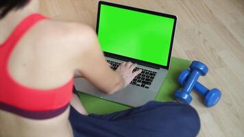 A woman in a sports bra and shorts using a laptop with a green screen ideal for app design digital content creation and fitness related mockup presentations video