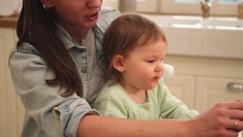 Happy family at home. Mother feeding her baby girl from spoon in kitchen. Little toddler child with messy funny face eats healthy food at home. Young woman mom giving food to kid daughter video