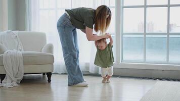 Baby development. First steps. Little infant girl making first steps at home. Happy mother supporting baby first steps. Mom helping toddler child walking in living room. Happy family at home video