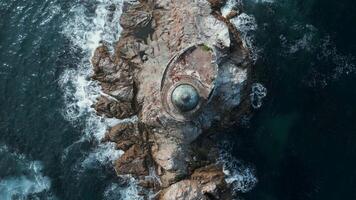 Top view of lighthouse on sea stone. Clip. Abandoned lighthouse on edge of rocky promontory in sea. Blue waves beat against stone ledge with lighthouse on sunny day video