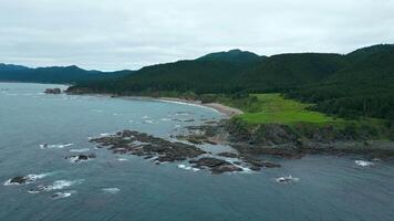 Top view of stone reefs off coast with green forests and rocks. Clip.Beautiful waves at reef shore with green forests of mountains. Northern landscape of green mountains and rocks on shore on cloudy video