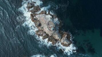 Top view of beautiful reef with waves and lighthouse. Clip. Single lighthouse on rocky ledge in blue sea on sunny day. Cinematic landscape of abandoned lighthouse on rocks in fantastic sea video