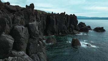 Top view of black rocks with blue waves on cloudy day. Clip. Black stone cliffs on ocean shore on cloudy day. Northern coasts on cloudy summer days video