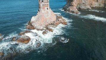 Top view of abandoned lighthouse in sea on sunny day. Clip. Abandoned lighthouse on rocks with waves of blue sea. Beautiful lighthouse on background of sea waves crashing on rocks video