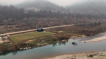 Aerial view Riverside cabin with fenced yard in a misty valley, A small black cabin with a fenced yard sits by a calm river in a misty valley video