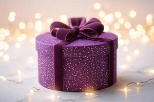 Purple round gift box with velvet bow on white table with fairy lights creating bokeh effect photo
