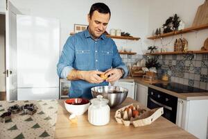 A young handsome man cooks in the kitchen at home. An attractive man breaks eggs into a metal bowl. Plates, a pot of sugar and a cookie jar are on the table. photo