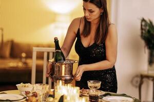 An attractive brunette in an evening dress puts a bucket of ice cubes and a bottle of champagne on the table. A young charming woman at a party. photo