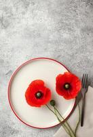 Red poppy flowers on a white plate with fork and napkin on a textured gray surface photo