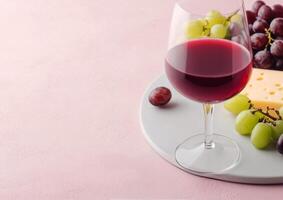 A glass of red wine beside a cheese platter with grapes on a pink table photo
