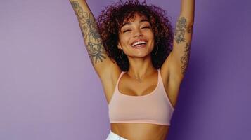 Joyful woman with curly hair and tattoos posing against a purple background during daytime photo