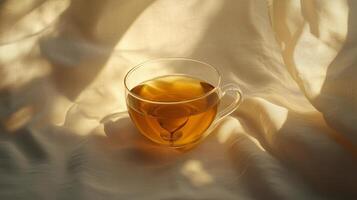 A clear glass cup of golden tea reflects sunlight on a soft tablecloth in a serene setting photo