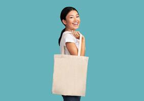 Smiling Asian Girl Holding White Eco Tote Bag Smiling Standing Over Turquoise Studio Background. Ecology And Eco-Friendly Lifestyle Concept. photo