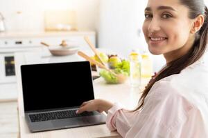 Portrait of happy smiling young lady using personal computer with black empty screen for mock up design, looking back sitting at dinner table in kitchen, working and typing on keyboard photo