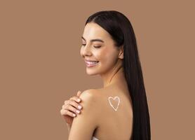 Smiling attractive shirtless long-haired brunette young woman with white heart-shaped symbol made of body lotion or cream on shoulder touching her smooth skin, isolated on beige studio background photo