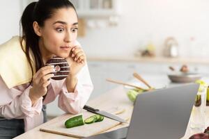 Reaction Concept. Terrified young woman watching reality show or news at home, using laptop device, eating sweet snack, leaning on kitchen counter while cooking tasty salad for dinner photo