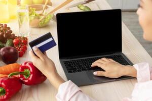 Webshop Concept. Over the shoulder view of unrecognizable positive woman using laptop pc with empty black screen for mock up, sitting at kitchen counter, holding and showing credit card, paying online photo