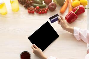 Webshop And E-commerce Concept. Top above high angle view of woman using tablet with empty black screen for mock up, sitting at kitchen counter, holding credit card, paying online, drinking coffee photo