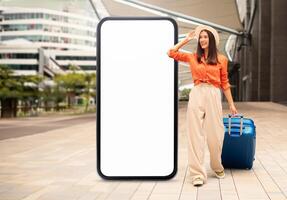 A traveler in a stylish outfit stands beside a gigantic blank smartphone display, waving while a blue suitcase rests beside her. The modern architecture of the location adds to the scene's appeal. photo