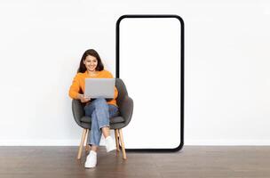 A woman in an orange sweater is happily using her laptop while seated in a modern chair next to large phone with blank screen, showcasing a relaxed atmosphere in a contemporary home setting. photo