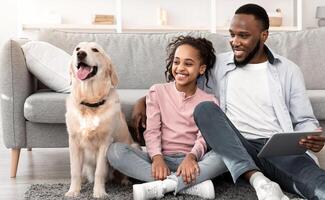 Lifestyle Concept. Portrait of smiling black man using digital tablet, sitting on the floor carpet and embracing daughter, looking at happy dog. Family spending time at home, having fun photo