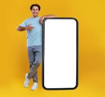 Check This Website. Smiling male leaning on smart phone with white blank screen, showing device and pointing finger at gadget with empty free copy space for mock up, recommending app, full body length photo