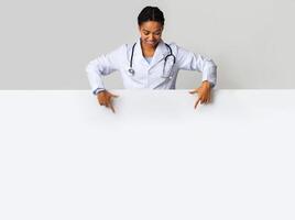 Medical offer. Afro female doctor looking down and pointing at blank white advertising board with empty space for text photo