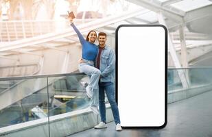 A cheerful couple poses joyfully beside a large blank screen in a modern indoor space. The woman raises her phone, while the man stands beside her with a smile, showcasing a lively moment. photo