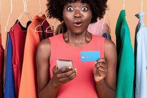 Mobile Shopping Application. Amazed Black Female Holding Cellphone And Credit Card Looking At Camera In Amazement Posing Near Clothing Rack Purchasing Clothes Online On Yellow Background In Studio photo