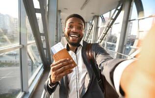Happy African Businessman Traveler Making Selfie With Passport And Flight Tickets In Airport Indoors. Celebrating End Of Lockdown. Selective Focus photo