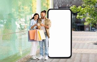 A happy couple stands outside a store next to huge phone with blank screen, looking at their phone and smiling. They hold colorful shopping bags, enjoying their day together under clear skies. photo