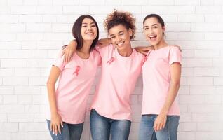 Happy Diverse Ladies In Pink Breast Cancer T-Shirts Hugging Posing Over White Brick Wall Background. Oncology Awareness Concept photo