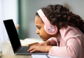 Youth and technology concept. Focused black schoolgirl using laptop computer, sitting at table too close to pc, side view. Little girl with poor eyesight having near vision problem photo