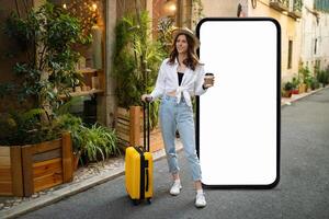 A traveler dressed in a stylish outfit holds a coffee cup while standing beside a bright yellow suitcase. The person poses in a charming street surrounded by greenery and buildings. photo