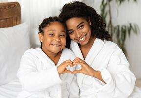 Happy Black Mom And Daughter In Bathrobes Making Heart Sign Form Hands, Connecting Their Arms, Having Fun And Bonding At Home, Relaxing On Bed Together After Bath, Smiling At Camera, Closeup photo