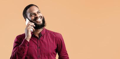 Phone Talk. Portrait Of Happy African Guy Chatting On Cellphone Standing Over Yellow Studio Background, Isolated. Smiling Black Man Speaking On Mobile Gadget, Looking Up, Copy Space photo