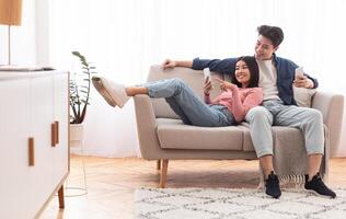 Asian Millennial Couple Using Smartphones Relaxing Sitting On Couch At Home On Weekend. Gadget Lifestyle, Mobile Application, Phone Users Browsing Internet Concept. Empty Space photo