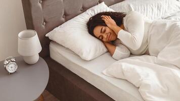 Portrait of irritated young black lady lying in bed and covering ears with hands, hearing and suffering from too loud sound or noise. Woman cannot sleep, doesn't want to wake up in the morning photo