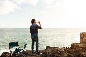 A man stands on a rocky cliff, facing the ocean and sipping a drink, as the sun begins to set. A nearby folding chair adds to the serene atmosphere, creating a sense of calmness and leisure. photo