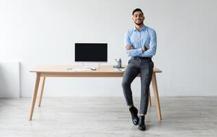 Mock Up Template. Happy Smiling Arab Man Sitting On Desk Posing With Folded Arms Showing Pc Computer With Blank Black Screen At Home Office, Free Copy Space. People, Technology, Remote Work Concept photo