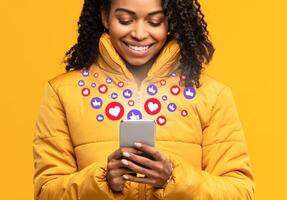 Happy Black Woman Holding Smartphone With Hearts And Like Buttons Standing On Yellow Studio Background. Phone User Networking Online Using Social Media, Reading Feed News, Commenting And Sharing Posts photo