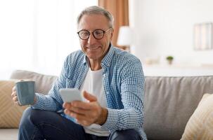 Portrait of smiling mature man in eyeglasses using mobile phone, watching and holding cup of coffee, sitting on the couch in living room. Guy browsing internet, surfing web, free copy space photo