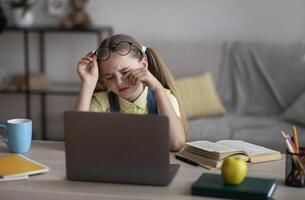Tired Student. Portrait of kid taking off glasses, touching dry eyes, sitting at table working on laptop photo