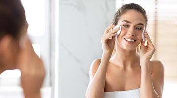 mujer utilizando algodón almohadillas limpieza cara con micelar agua cuidando para piel mirando a espejo en pie en moderno baño a hogar. joven dama disfrutando facial protección de la piel rutina concepto. panorama foto